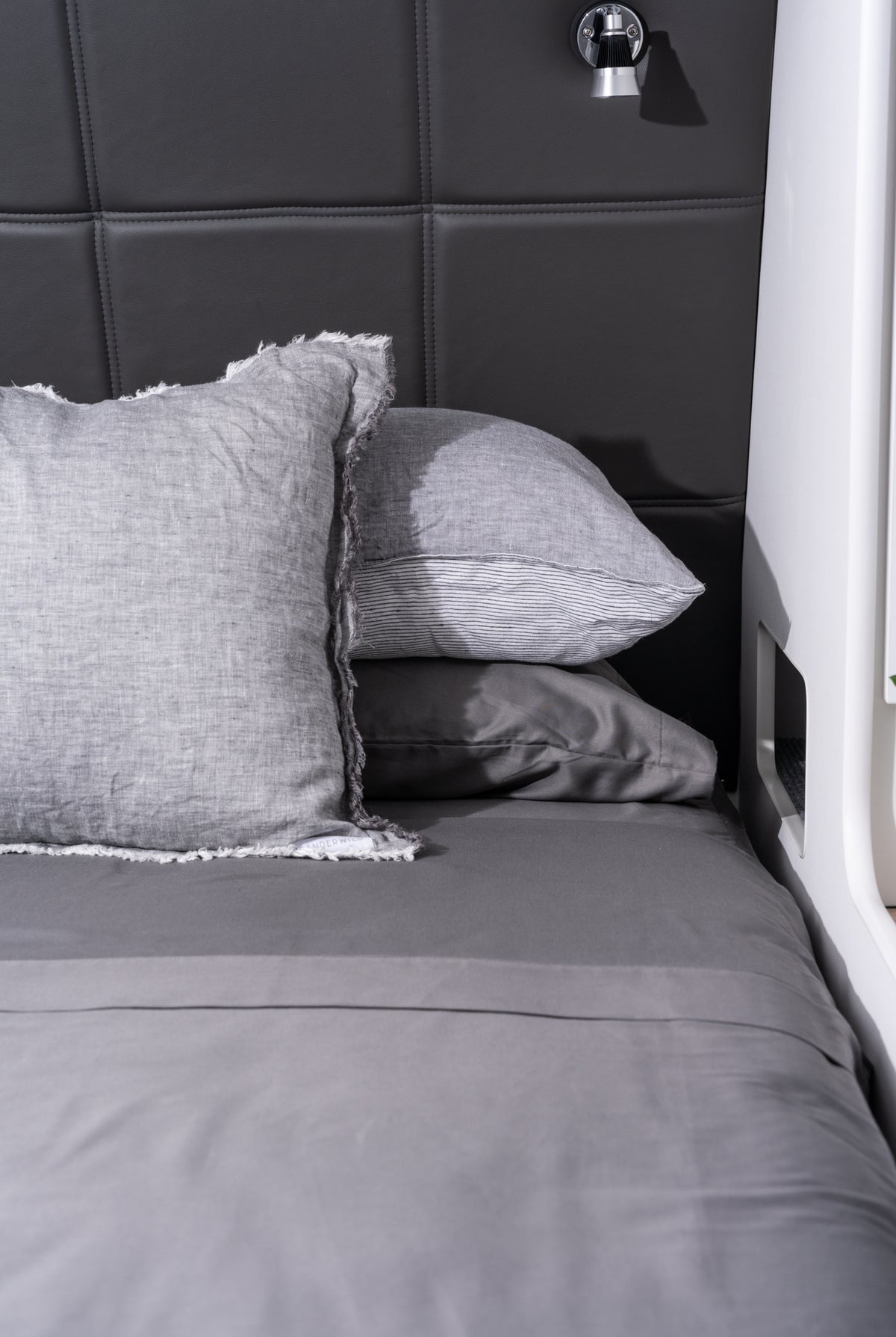 A close-up of a bed with gray bedding, including a textured gray pillow with frayed edges, a smooth gray pillow, and a matching gray duvet, set against a dark gray padded headboard.