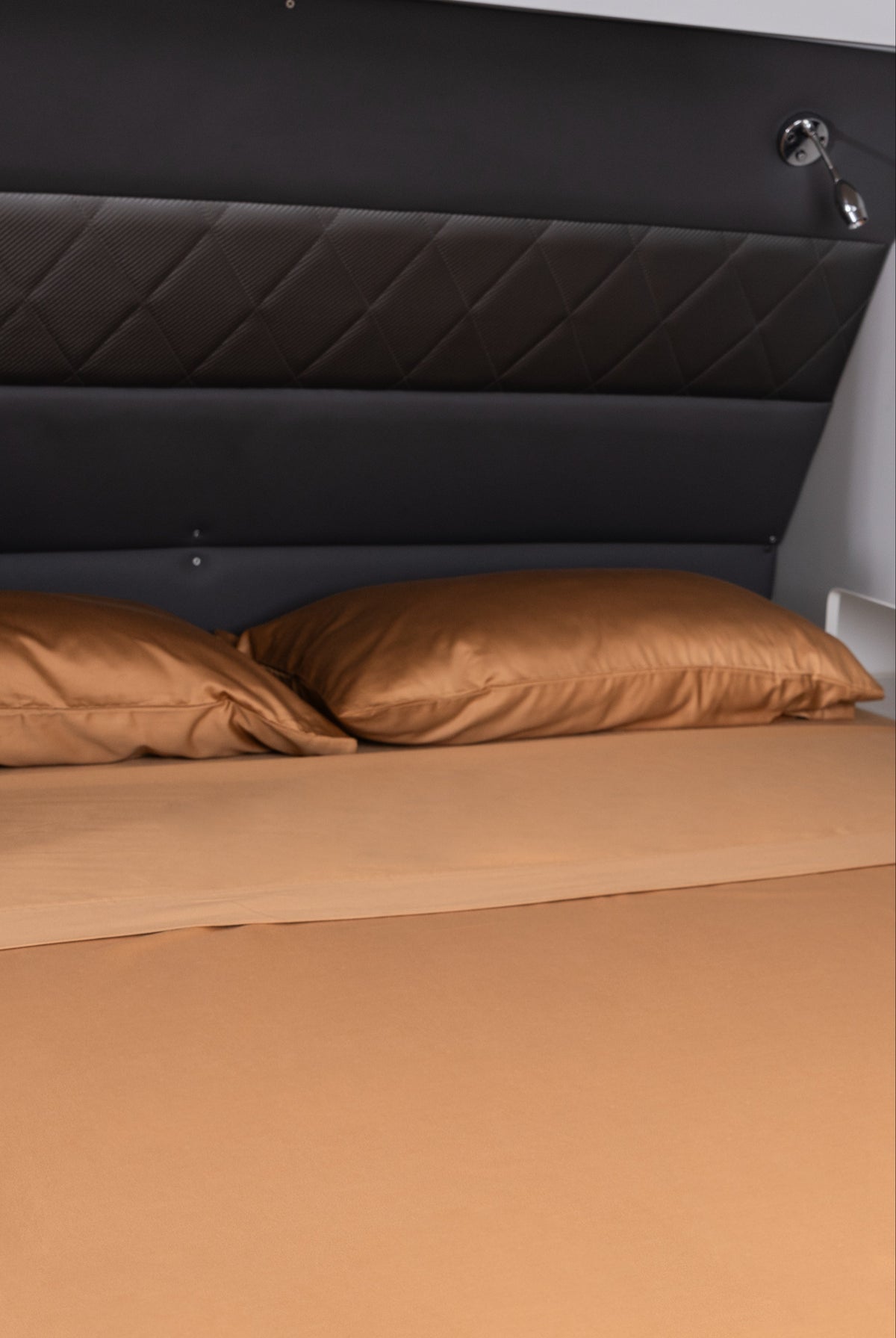 A fitted sheet in a warm tan or camel color covers a bed, with two matching pillowcases and a dark padded headboard behind it.