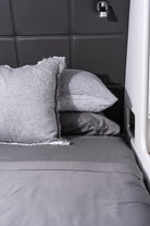 A close-up of a bed with gray bedding, including a textured gray pillow with frayed edges, a smooth gray pillow, and a matching gray duvet, set against a dark gray padded headboard.