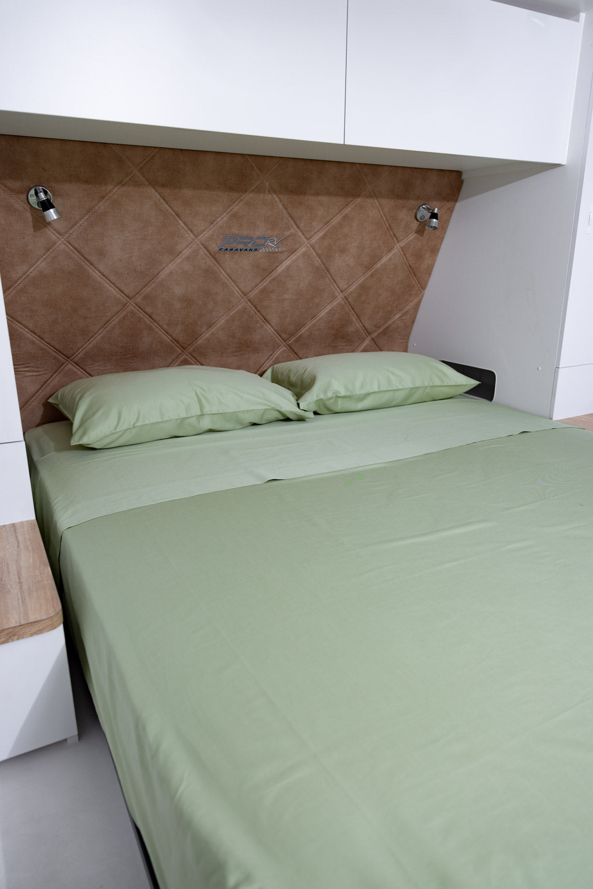 A bed dressed in light sage green bedding, positioned against a brown diamond-quilted headboard with white wall cabinets above.
