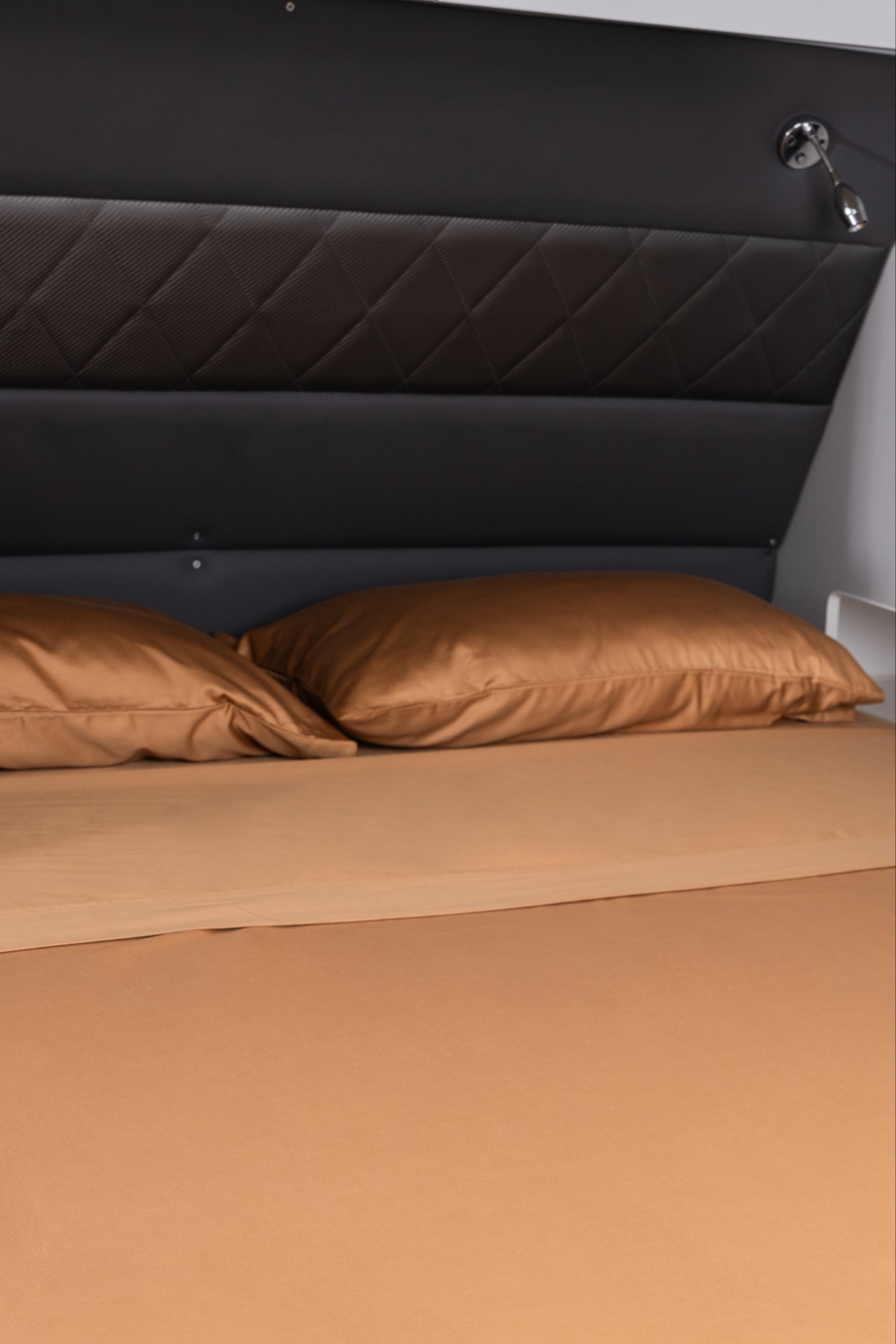 A fitted sheet in a warm tan or camel color covers a bed, with two matching pillowcases and a dark padded headboard behind it.