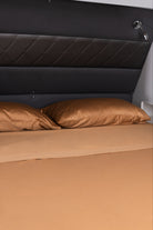 A fitted sheet in a warm tan or camel color covers a bed, with two matching pillowcases and a dark padded headboard behind it.